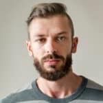 Młody mężczyzna z poważnym wyrazem twarzy, krótkimi brązowymi włosami ułożonymi na bok i pełną brodą, ubrany w szaro-zieloną koszulę w paski, stoi na prostym jasnym tle - idealnym do reprezentowania usług pomocy drogowej lub lawety.