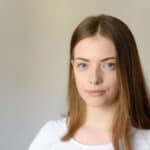 Młoda kobieta o prostych, jasnobrązowych włosach i niebieskich oczach, ubrana w biały top, stoi przed prostym, jasnym tłem, spokojnie patrząc w kamerę, jakby czekała na pomoc drogową.