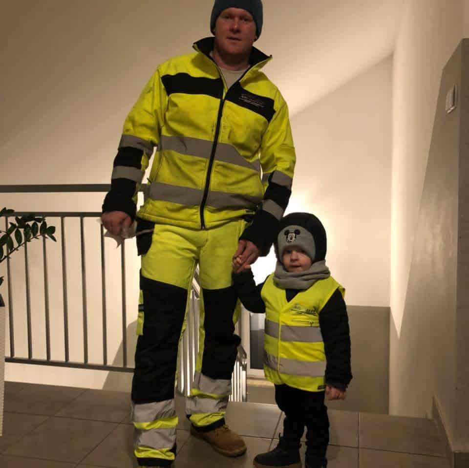 Dorosły i dziecko stoją w pomieszczeniu, oboje w dopasowanych żółtych strojach roboczych o wysokiej widoczności, często spotykanych w zespołach pomocy drogowej, trzymając się za ręce. Dziecko nosi również czapkę beanie z uszami niedźwiedzia. Znajdują się w pobliżu schodów z białymi poręczami.