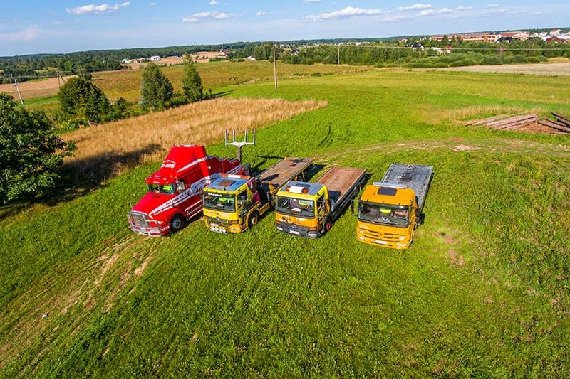 Widok z lotu ptaka na cztery ciężarówki, w tym czerwoną lawetę i trzy żółte pojazdy pomocy drogowej, zaparkowane obok siebie na trawiastym polu z drzewami, kłodami i polami uprawnymi w tle.