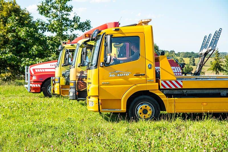Trzy lawety, dwie żółte i jedna czerwona, zaparkowane na trawiastym polu pod słońcem, gotowe do pomocy drogowej i holowania, z drzewami i otwartym krajobrazem w tle.