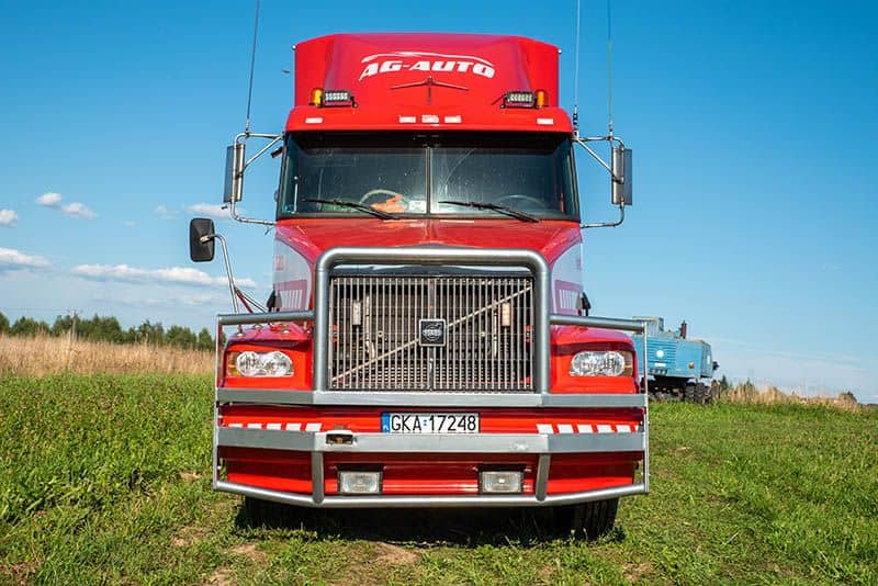 Jasnoczerwona ciężarówka Volvo ze znakiem "AG-AUTO" pomoc drogowa na górze jest zaparkowana na trawie, przodem do kierunku jazdy. Inna niebieska ciężarówka jest widoczna w tle pod czystym, błękitnym niebem.