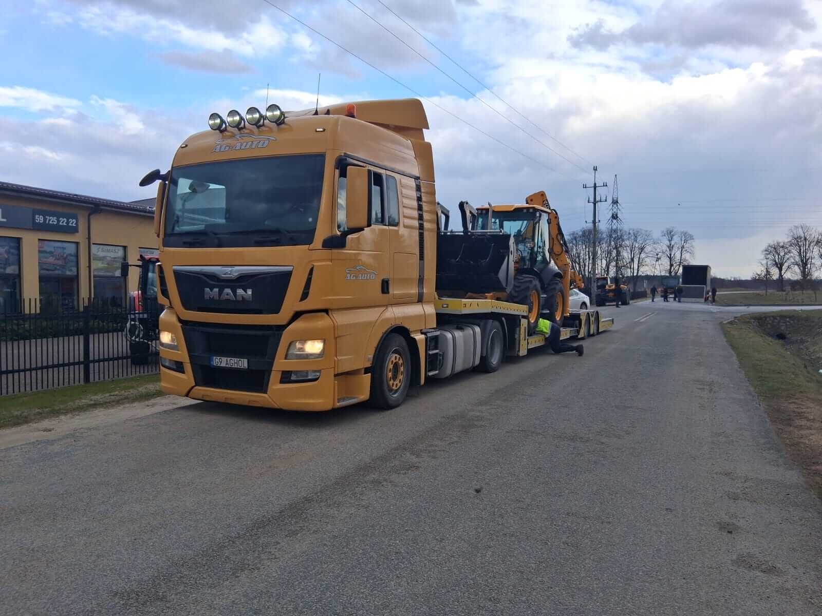 Żółta ciężarówka MAN z przyczepą zaparkowana na wiejskiej drodze, przewożąca koparko-ładowarkę w ramach usługi lawety. W tle znajdują się budynki, drzewa i inne pojazdy pod częściowo zachmurzonym niebem.