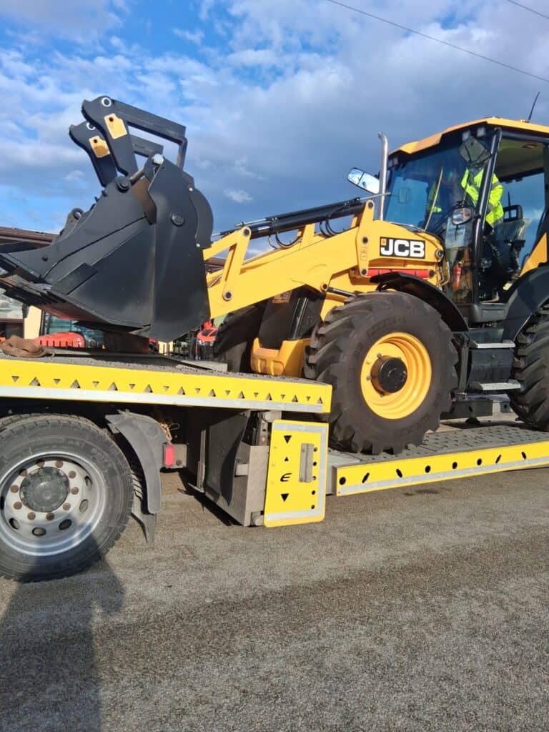 Żółta koparko-ładowarka JCB jest transportowana na lawecie pod częściowo zachmurzonym niebem. Duża przednia łyżka i kabina ładowarki są dobrze widoczne, gotowe do szybkiej pomocy drogowej lub holowania.