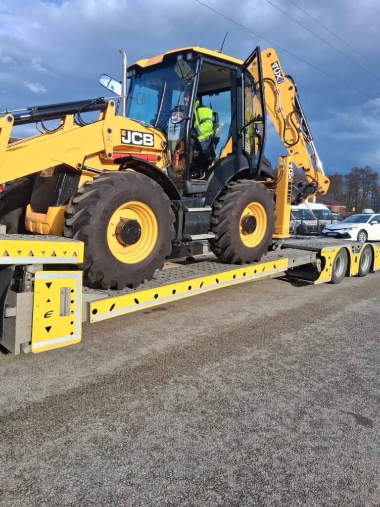 Żółta koparko-ładowarka JCB jest transportowana na żółtej lawecie, prawdopodobnie do holowania. Pracownik w kamizelce odblaskowej siedzi wewnątrz kabiny, podczas gdy kilka pojazdów jest zaparkowanych w pobliżu w ten pochmurny dzień, co sugeruje pomoc drogową.