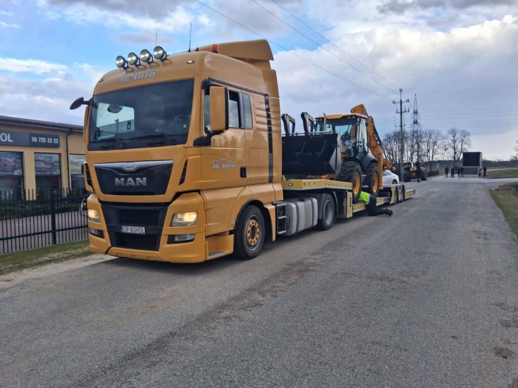 Żółta ciężarówka MAN, używana do lawety i holowania, jest zaparkowana na poboczu drogi z koparko-ładowarką na przyczepie. W tle widać budynki, ogrodzenie i zachmurzone niebo.