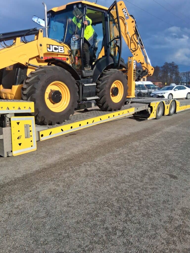 Żółta koparko-ładowarka JCB jest ładowana na lawetę na utwardzonej nawierzchni. Osoba w kamizelce odblaskowej obsługuje pojazd, sugerując pomoc drogową. W tle kilka samochodów zaparkowanych pod zachmurzonym niebem.