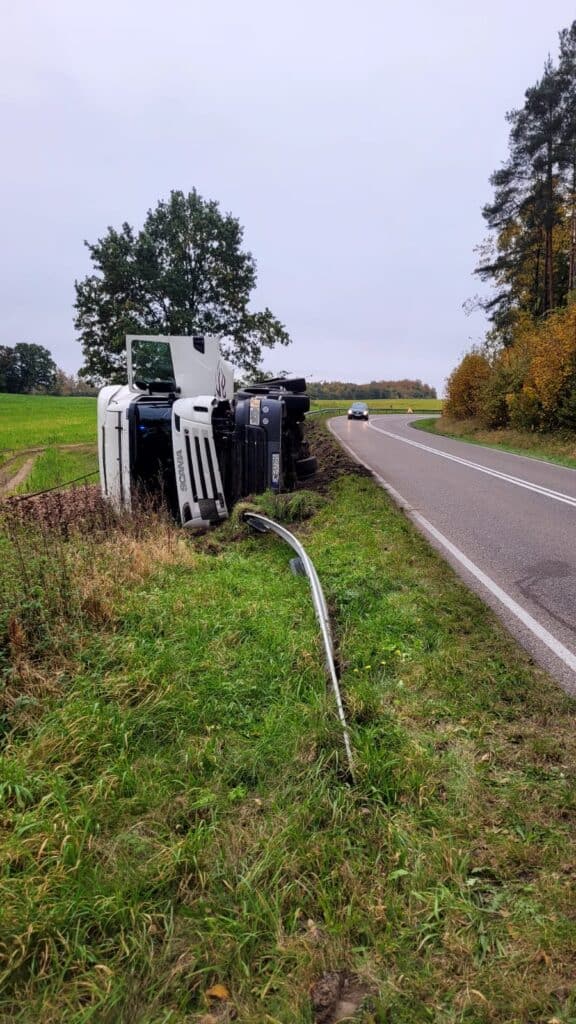 Biała ciężarówka przewróciła się na bok w trawiastym rowie obok zakrzywionej wiejskiej drogi, z wygiętą barierką ochronną na pierwszym planie i samochodami jadącymi obok - idealne miejsce na pomoc drogową lub pomoc lawety na miejscu zdarzenia.