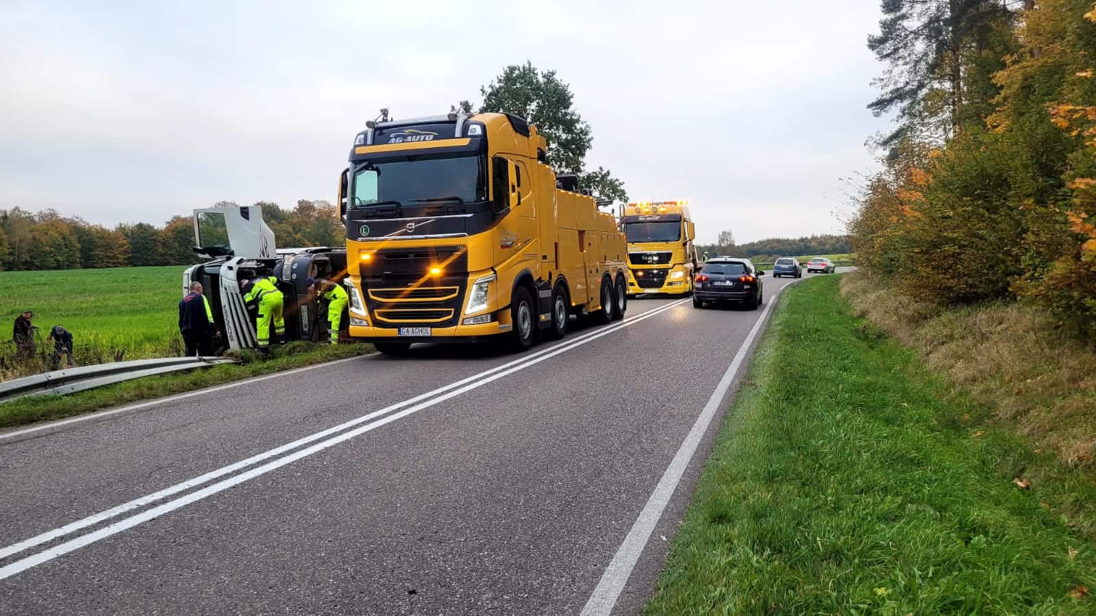 Dwie duże żółte lawety holownicze są zaparkowane na wiejskiej drodze w pobliżu przewróconego pojazdu, podczas gdy ludzie w kamizelkach bezpieczeństwa pracują w pobliżu, a samochody przejeżdżają obok. Wokół zielone pola i drzewa.