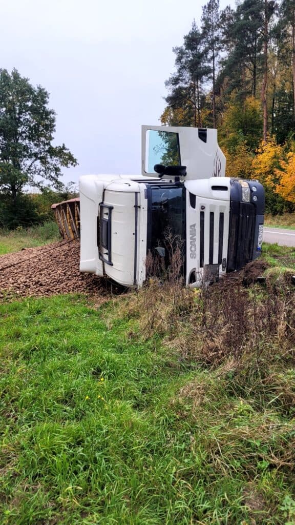 Duża biała ciężarówka leży przewrócona na bok przy wiejskiej drodze, a jej ładunek żwiru rozsypał się na trawę. Wysokie drzewa z jesiennymi liśćmi są w tle, gdy laweta przybywa, aby zapewnić pomoc drogową.