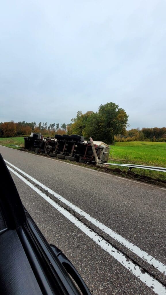 Przewrócona przyczepa leży przewrócona obok wiejskiej drogi z podwójnymi białymi liniami, obok zielonych pól i drzew pod zachmurzonym niebem. Scena czeka na pomoc drogową, widziana z przejeżdżającego pojazdu.