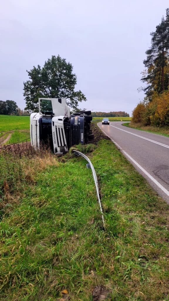 Duża biała ciężarówka przewróciła się na bok w trawiastym rowie obok wiejskiej drogi, w pobliżu uszkodzonej barierki ochronnej. Samochód odjeżdża, podczas gdy laweta jest potrzebna do holowania i pomocy drogowej wśród zielonych pól i drzew.