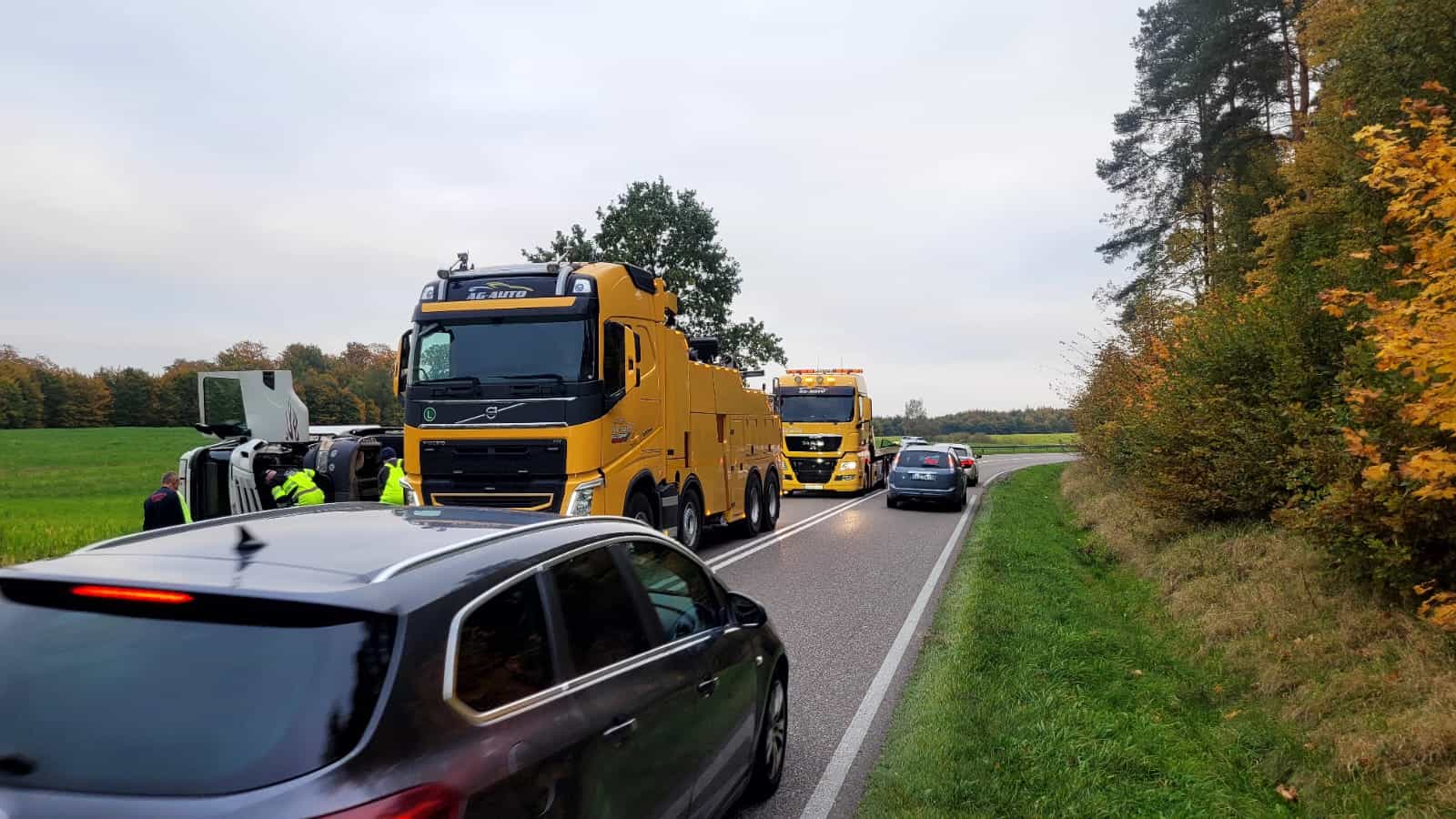 Kilka dużych żółtych lawet pomaga na miejscu wypadku samochodowego na wiejskiej drodze. Czarny samochód przejeżdża obok, podczas gdy pomoc drogowa w kamizelkach bezpieczeństwa stoi w pobliżu przewróconego pojazdu. Wzdłuż drogi rosną drzewa i pola.