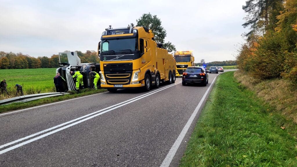 Kilka żółtych lawet zapewnia pomoc drogową, pomagając przy przewróconym pojeździe na poboczu wiejskiej drogi. Samochody policyjne i pracownicy są obecni, ruch jest częściowo zablokowany, a drzewa i pola otaczają miejsce zdarzenia pod zachmurzonym niebem.