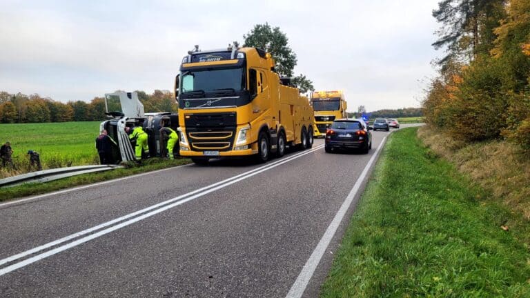 Kilka żółtych lawet zapewnia pomoc drogową, pomagając przy przewróconym pojeździe na poboczu wiejskiej drogi. Samochody policyjne i pracownicy są obecni, ruch jest częściowo zablokowany, a drzewa i pola otaczają miejsce zdarzenia pod zachmurzonym niebem.