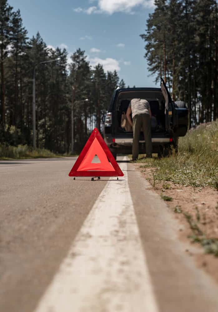Czerwony trójkąt ostrzegawczy umieszczony na poboczu drogi, z osobą stojącą przy otwartym bagażniku czarnego samochodu, prawdopodobnie czekającą na pomoc drogową. Scena rozgrywa się na wiejskiej drodze otoczonej wysokimi drzewami.