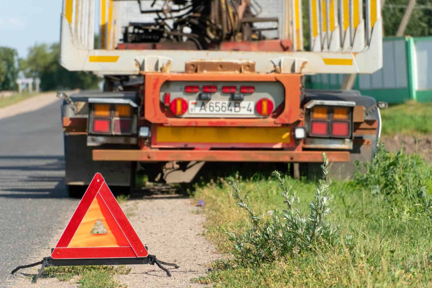 Czerwony trójkąt ostrzegawczy jest umieszczony na poboczu drogi w pobliżu tyłu dużej zatrzymanej ciężarówki, sygnalizując awarię na drodze. Pomoc drogowa lub laweta może być potrzebna na tej wiejskiej drodze porośniętej trawą i roślinami.