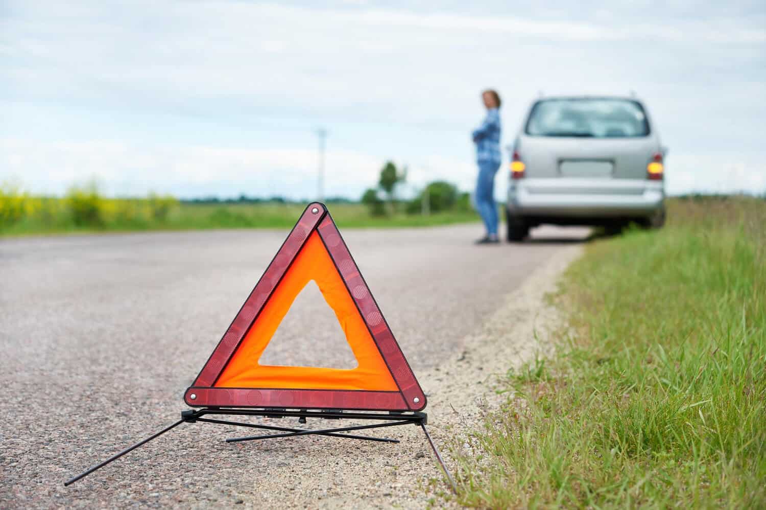 Trójkąt ostrzegawczy umieszczony na poboczu wiejskiej drogi. W tle osoba stoi obok zaparkowanego samochodu, prawdopodobnie czekając na pomoc drogową lub holowanie po nagłym wypadku drogowym. Scena rozgrywa się na zewnątrz w ciągu dnia.