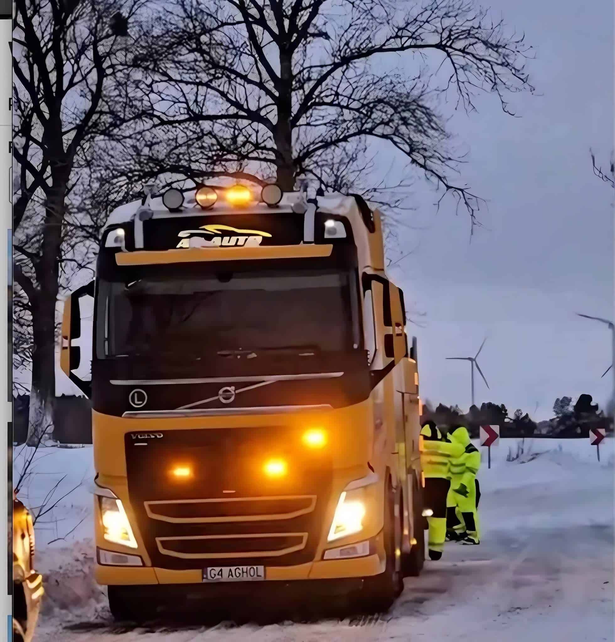 Duża żółta ciężarówka Volvo pomoc drogowa z migającymi światłami jest zaparkowana na zaśnieżonej drodze w pobliżu bezlistnych drzew. Dwie osoby w jaskrawożółtych odblaskowych ubraniach stoją obok ciężarówki, a w tle widoczne są turbiny wiatrowe.