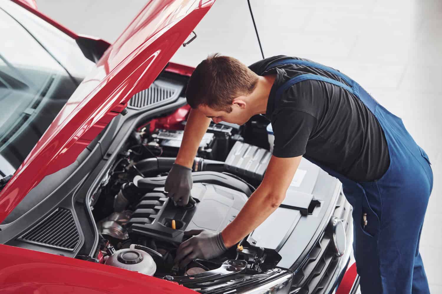 Mechanik w niebieskim kombinezonie i rękawiczkach sprawdza lub naprawia silnik czerwonego samochodu z otwartą maską w dobrze oświetlonym garażu, prawdopodobnie po przywiezieniu go przez pomoc drogową lub holowanie.