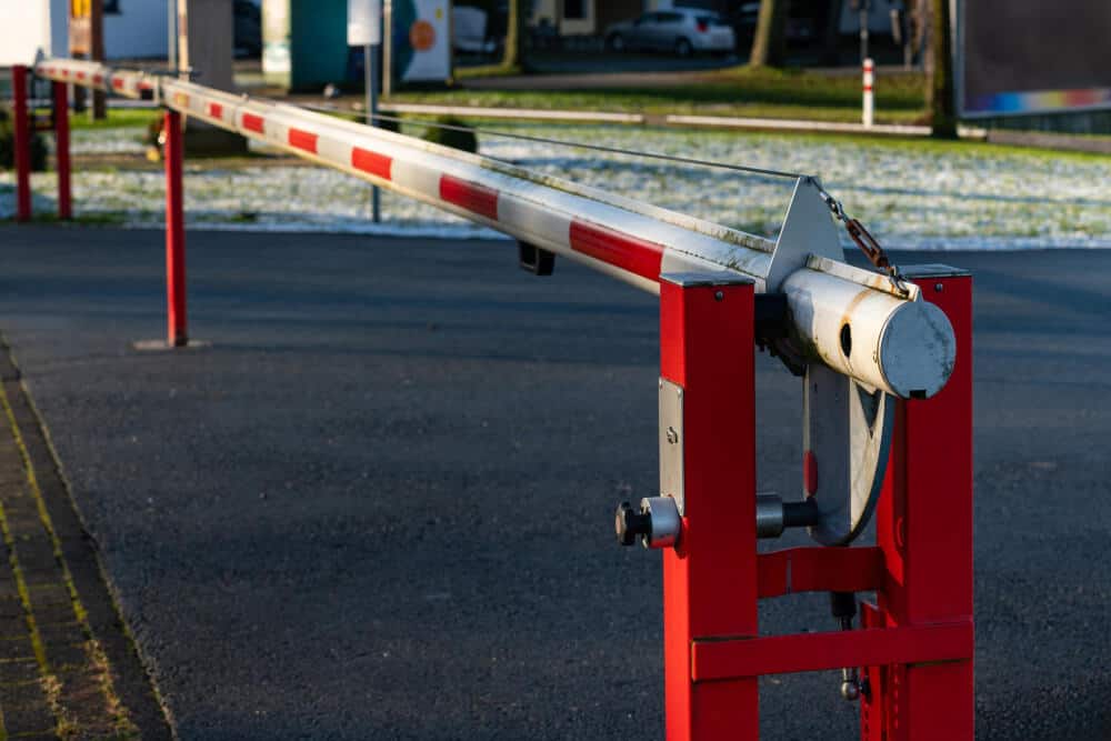 Biało-czerwony szlaban parkingowy blokuje utwardzony wjazd, a w tle w słoneczny dzień widać trawę, zaparkowane samochody i lawetę z serwisu pomoc drogowa.