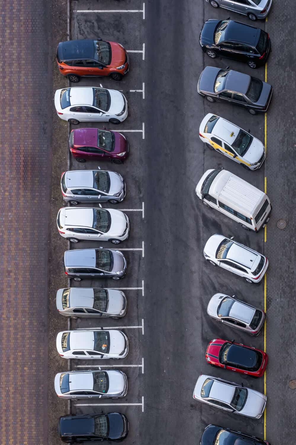 Widok z lotu ptaka na parking z samochodami zaparkowanymi w dwóch rzędach wzdłuż centralnego podjazdu. Po lewej stronie widoczna jest laweta, gotowa do holowania, wśród różnych samochodów zaparkowanych czołowo i równolegle, w różnych kolorach i rozmiarach.