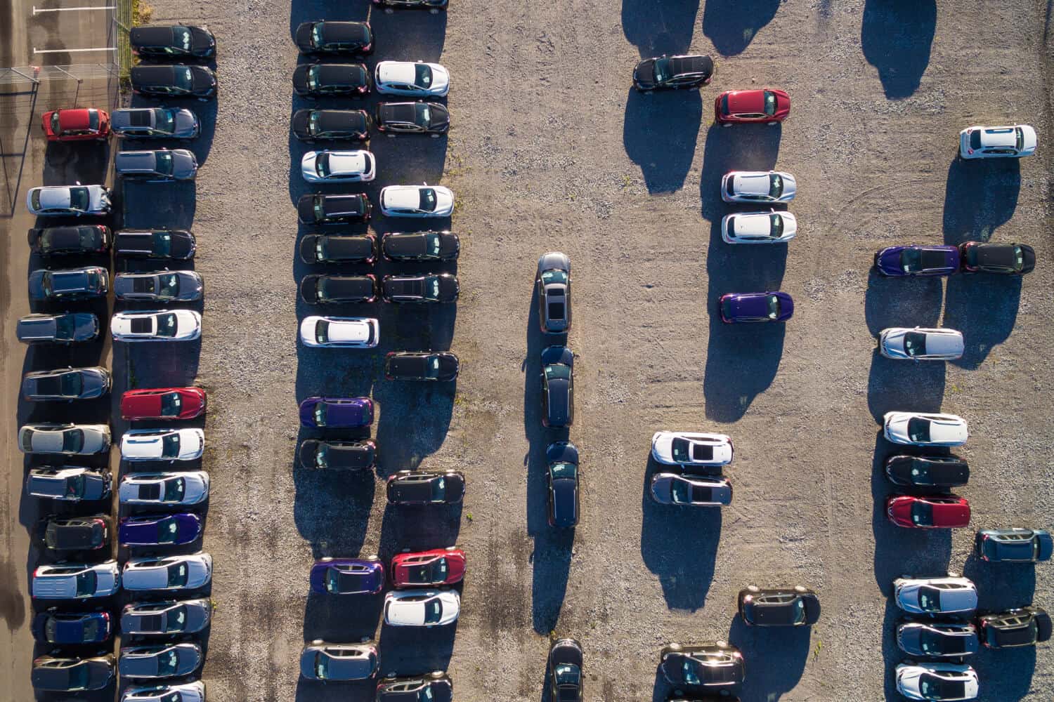 Widok z lotu ptaka na samochody zaparkowane w równych rzędach na żwirowym parkingu, z kilkoma pustymi miejscami parkingowymi i długimi cieniami pojazdów - wśród nich widoczna jest jedna laweta z lokalnej pomocy drogowej, gotowa w razie potrzeby pomóc w holowaniu.