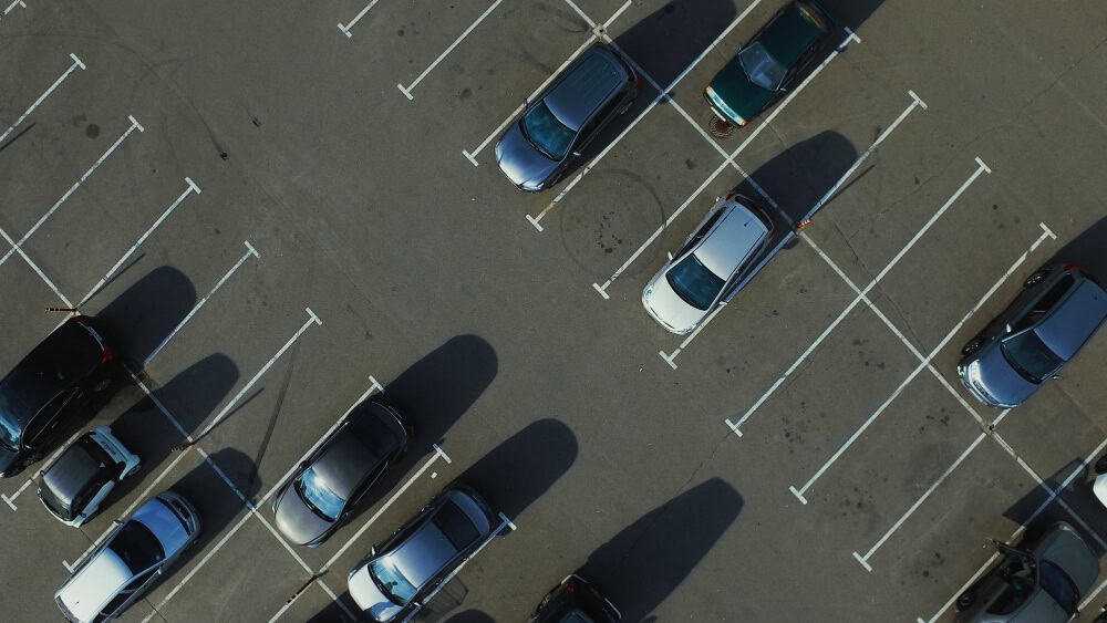 Widok z lotu ptaka na parking z kilkoma samochodami i kilkoma pustymi miejscami; cienie rozciągają się po chodniku. W pobliżu zaparkowana jest laweta, co sugeruje niedawne holowanie lub pomoc drogową.