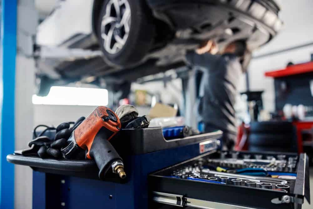 Mechanik pracuje pod samochodem podniesionym na podnośniku w warsztacie samochodowym, podczas gdy wózek narzędziowy zawiera różne narzędzia, rękawice i pneumatyczny klucz udarowy - gotowe do pomocy w razie potrzeby holowania lub pomocy drogowej.