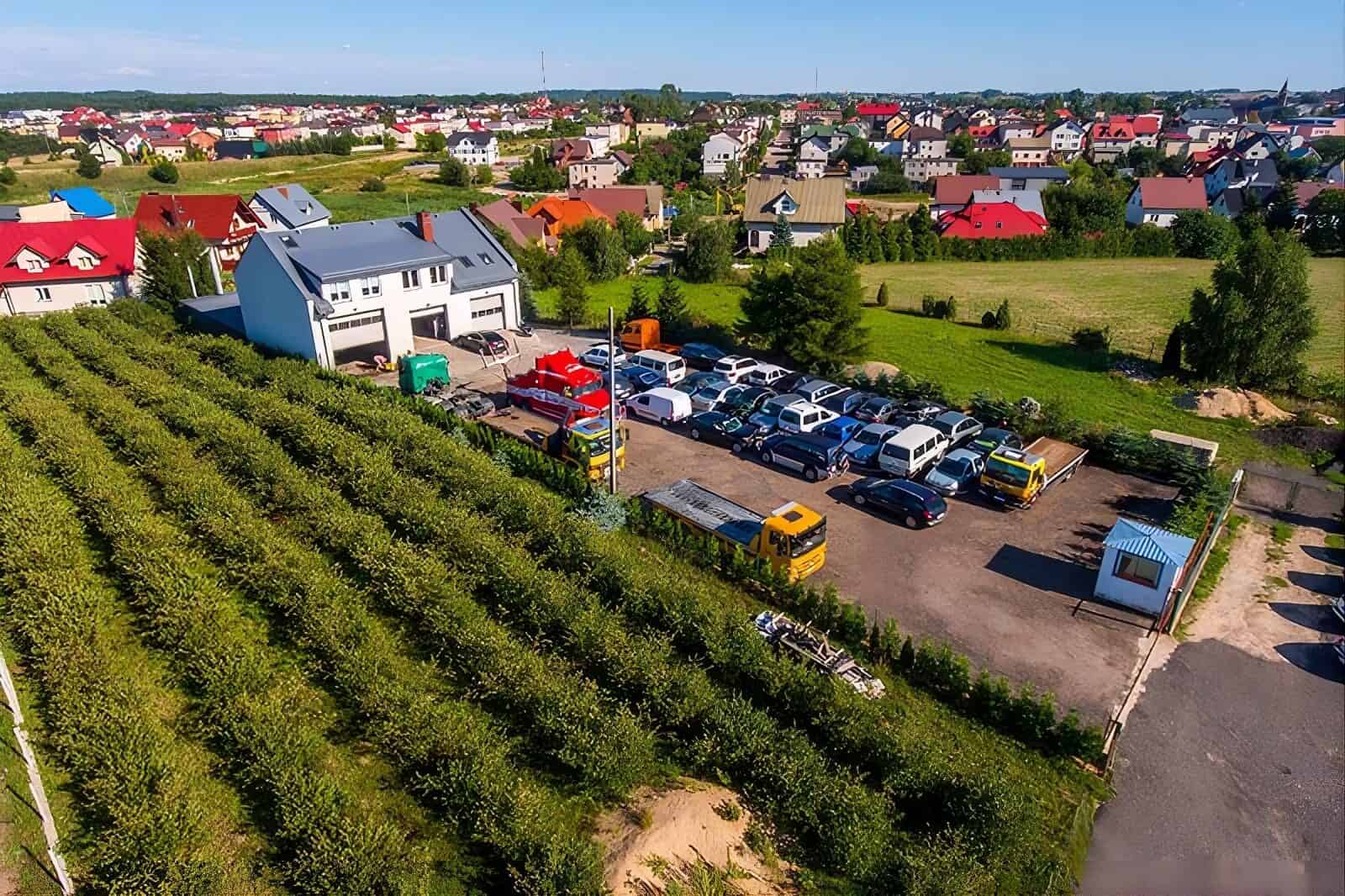 Widok z lotu ptaka na parking z kilkoma pojazdami, w tym lawetą, obok białego budynku, otoczony zielonymi żywopłotami i drzewami, z osiedlem kolorowych domów w tle.