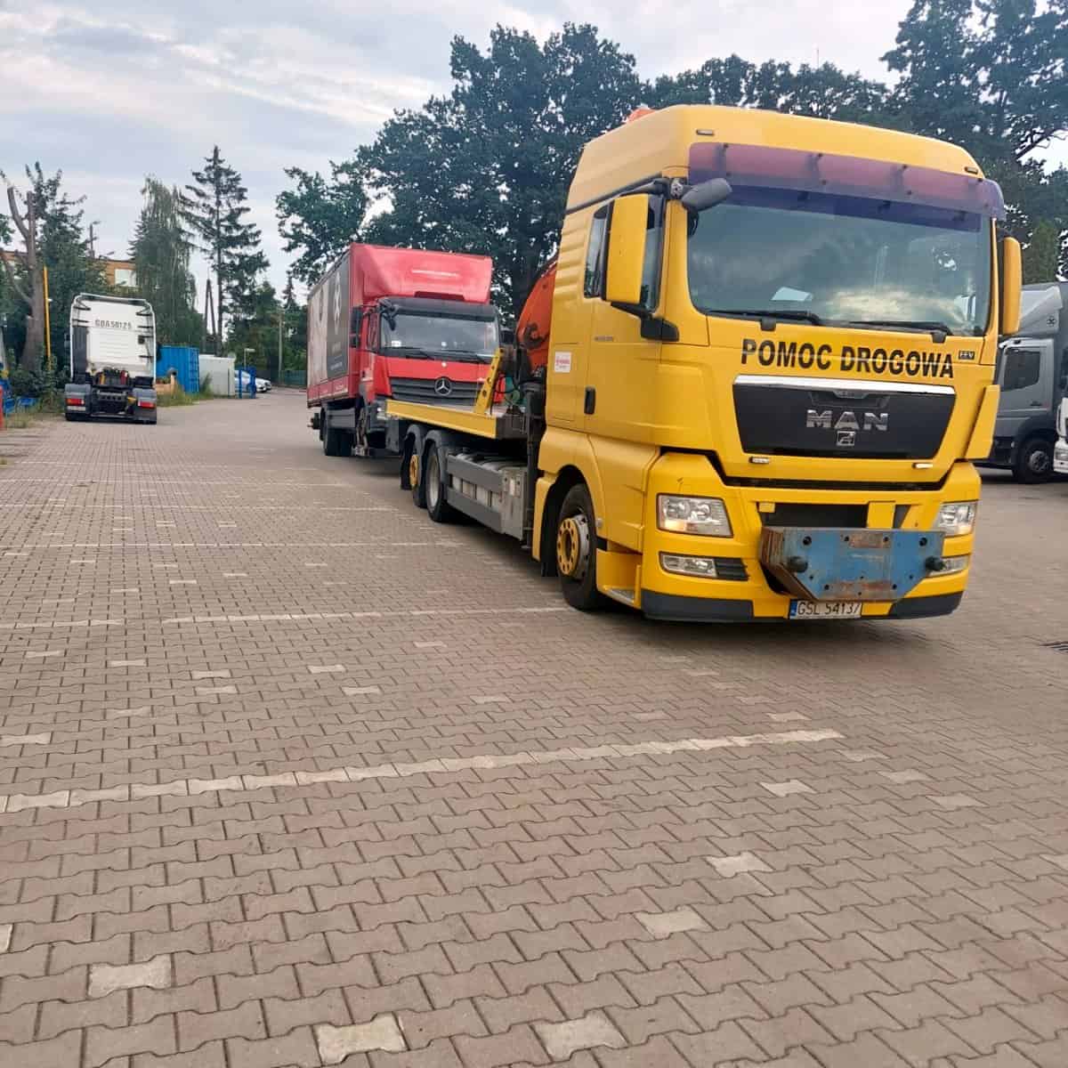 Żółta laweta z napisem "Pomoc Drogowa" przewozi czerwoną ciężarówkę na platformie na parkingu, w tle widoczne są inne pojazdy i drzewa.