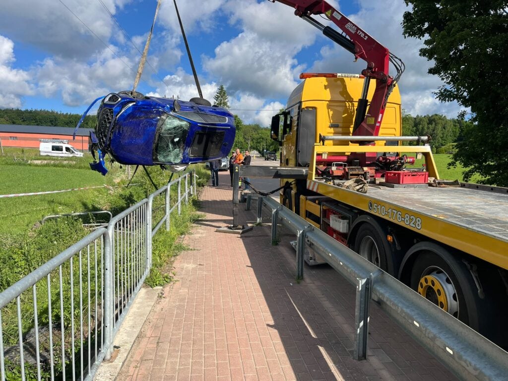 Laweta z dźwigiem podnosi poważnie uszkodzony niebieski samochód stojący przy drodze i chodniku z balustradami, a w tle, pod częściowo zachmurzonym niebem, widać zielone pola, galerię i drzewa.