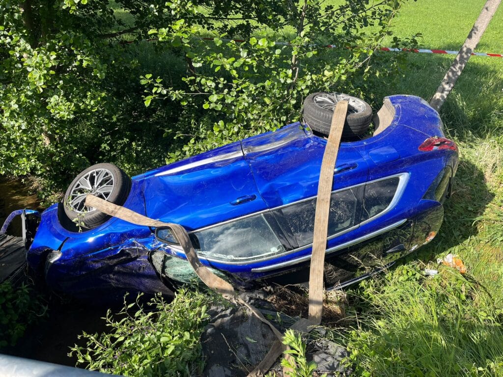 Niebieski samochód leży przewrócony na dach w zalesionym terenie porośniętym trawą. Pojazd wygląda na poważnie uszkodzony – brakuje mu jednego koła, a do karoserii przymocowane są pasy zabezpieczające. Jasne światło słoneczne oświetla tę scenę, nadając jej wygląd niczym z dramatycznej galerii.