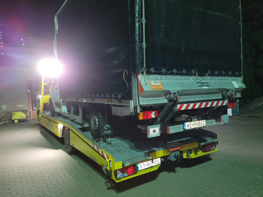 Żółta laweta przewozi w nocy przykrytą przyczepę na oświetlonym parkingu, tworząc scenę przypominającą galerię motoryzacyjną. Widoczne są tablica rejestracyjna i tylne światła przyczepy, a światła lawety jasno świecą.
