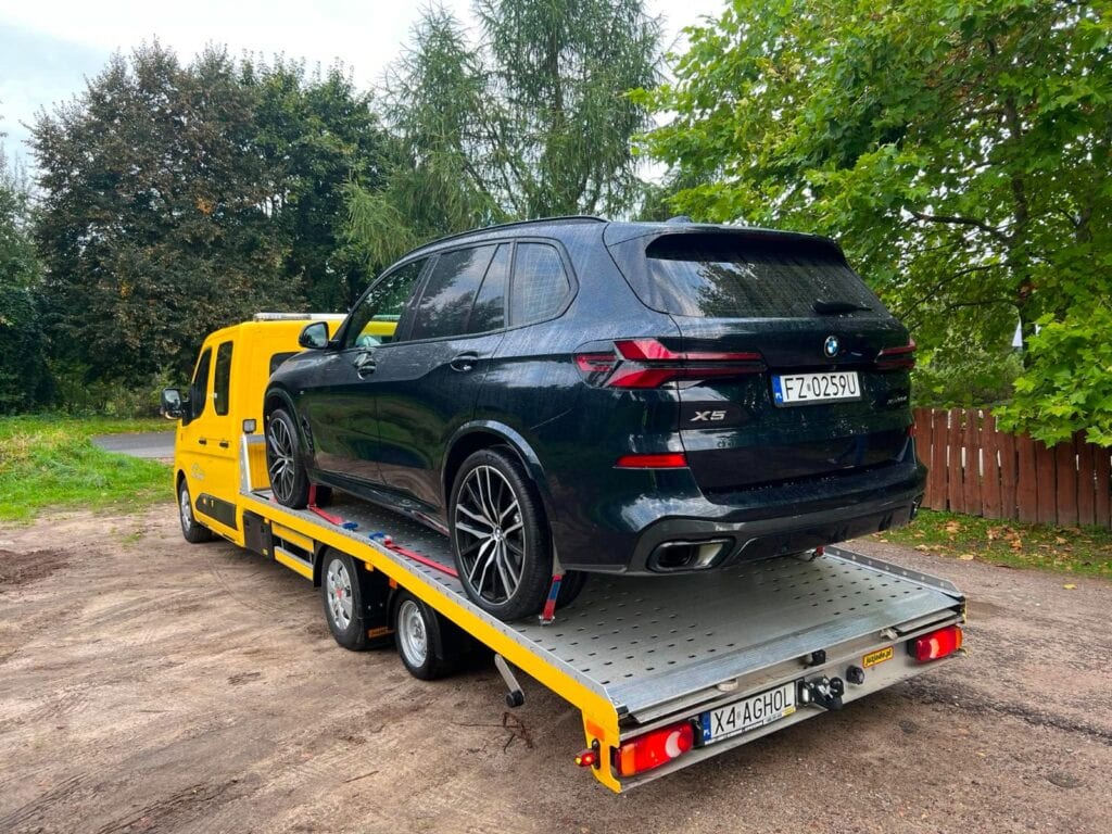 Żółta laweta przewozi na platformie czarnego SUV-a. Pojazdy stoją na świeżym powietrzu w pobliżu drzew i drewnianego ogrodzenia, tworząc scenę, która idealnie pasowałaby do galerii motoryzacyjnej.