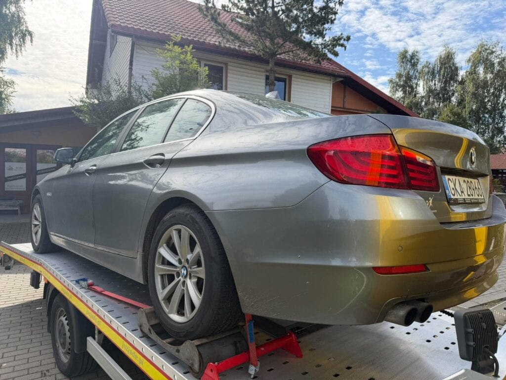 Srebrny sedan marki BMW stoi na platformie lawety w dzielnicy mieszkalnej, a w tle widać drzewa i dom, co tworzy scenę przypominającą ekspozycję w galerii motoryzacyjnej.
