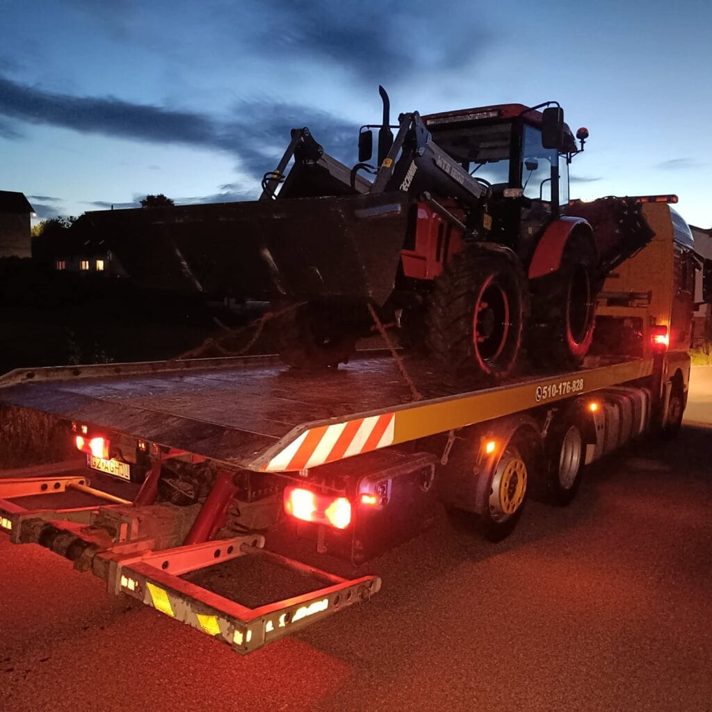 O zmierzchu czerwony traktor z ładowaczem czołowym jest zamocowany na platformie lawety, a jego światła lśnią na tle ciemniejącego nieba — to poruszający obraz, godny każdej galerii.