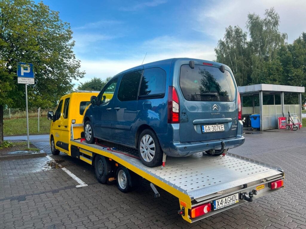 Na parkingu w pobliżu galerii handlowej niebieski samochód dostawczy marki Citroën jest ładowany na żółtą lawetę. W tle widać przystanek autobusowy i zaparkowane wózki sklepowe.