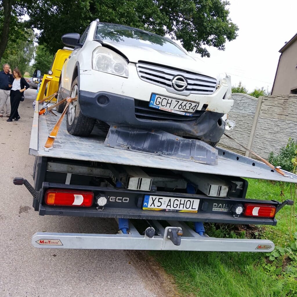 Uszkodzony biały samochód z wgniecionym przodem jest ładowany na lawetę z platformą na poboczu drogi; obok stoją dwie osoby, obserwujące tę scenę, jakby były częścią nieoczekiwanej wystawy.