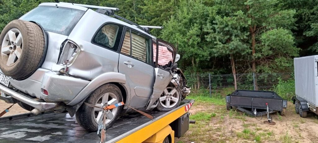 Uszkodzony srebrny SUV z wgniecionym tyłem i bokami stoi na żółtej lawecie, gotowy do przewiezienia do warsztatu. W tle widać drzewa i małą czarną przyczepę.