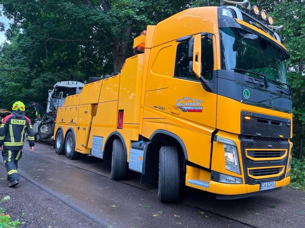 Duża żółta laweta stoi na mokrej drodze w pobliżu drzew, a strażak w stroju bojowym idzie w kierunku pojazdu holowanego za lawetą — obraz, który idealnie pasowałby do każdej galerii zdjęć przedstawiających dramatyczne akcje ratownicze na drogach.