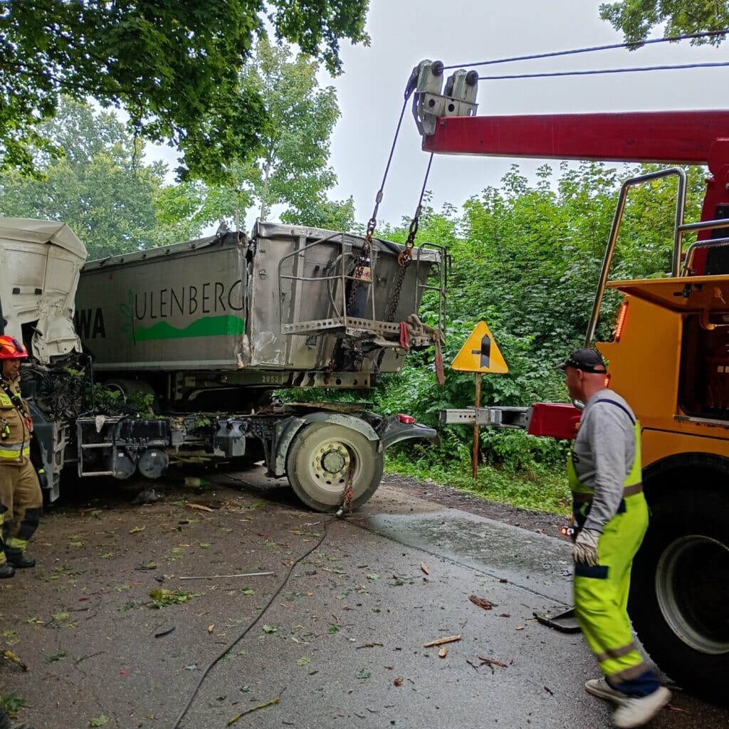 Laweta podnosi tył uszkodzonej ciężarówki w pobliżu leśnej drogi, a pracownicy w strojach ochronnych nadzorują tę operację, tworząc scenę godną każdej galerii poświęconej akcjom ratowniczym na drogach.