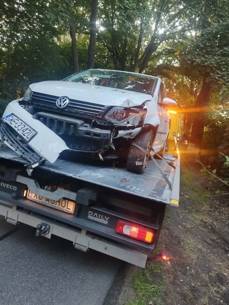 Srebrny Volkswagen z poważnymi uszkodzeniami przedniej części nadwozia jest ładowany na lawetę w zalesionym terenie. Zachodzące słońce tworzy efekt odblasku za lawetą, nadając tej scenie nastrojowy charakter przypominający artystyczną wystawę w galerii.