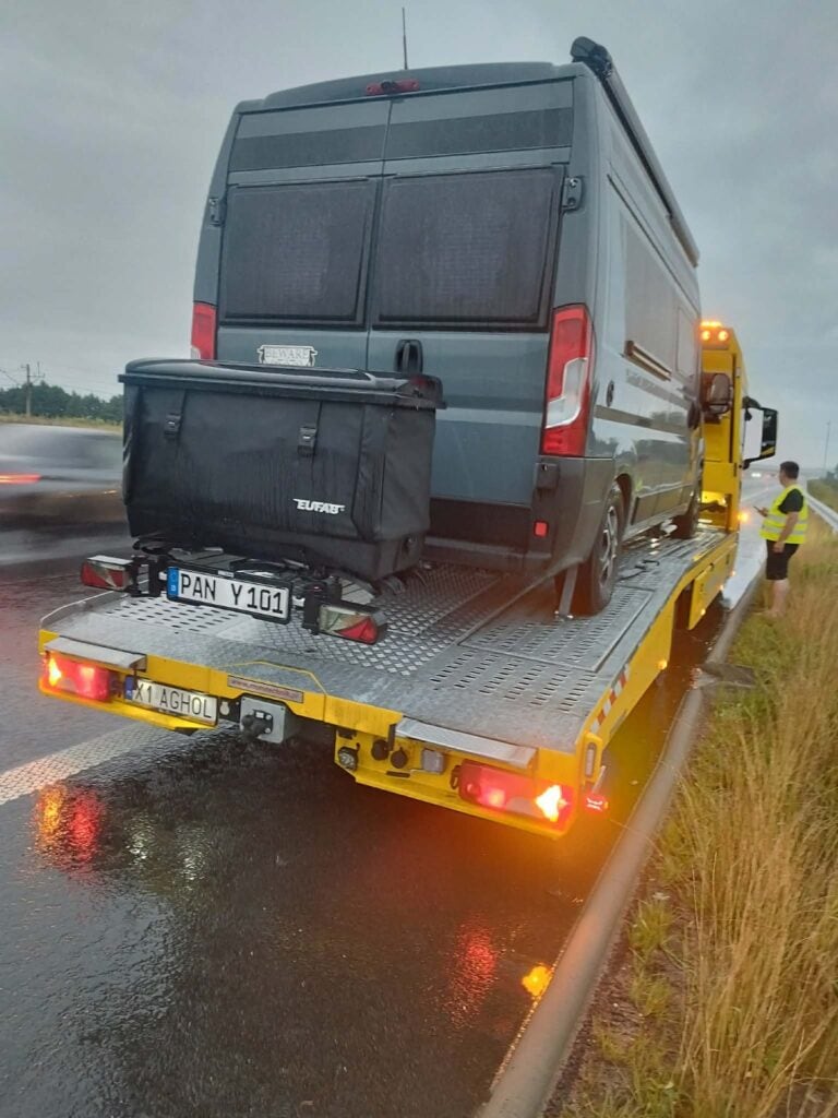 Czarny van z bagażnikiem dachowym jest ładowany na żółtą lawetę na mokrej drodze, tworząc scenę przypominającą motoryzacyjną galerię. W pobliżu stoi osoba w kamizelce odblaskowej, a w tle widać rozmyty w ruchu inny pojazd.