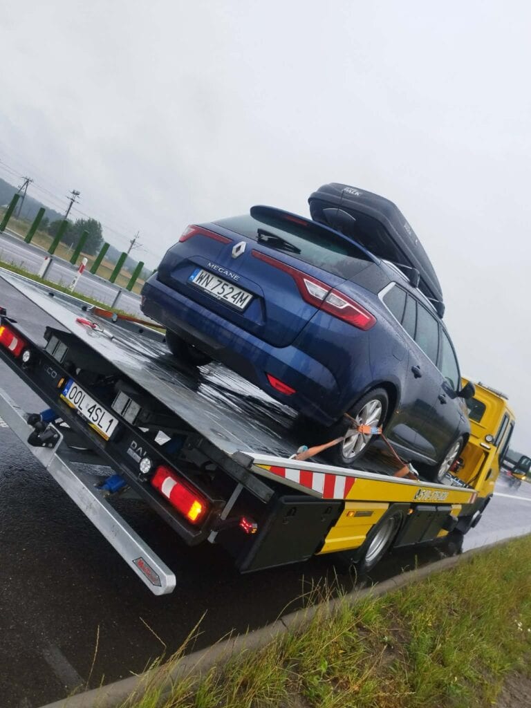 Niebieskie kombi marki Renault z bagażnikiem dachowym jest ładowane na żółtą lawetę na mokrej drodze podczas deszczowej pogody, tworząc scenę godną każdej galerii motoryzacyjnej.